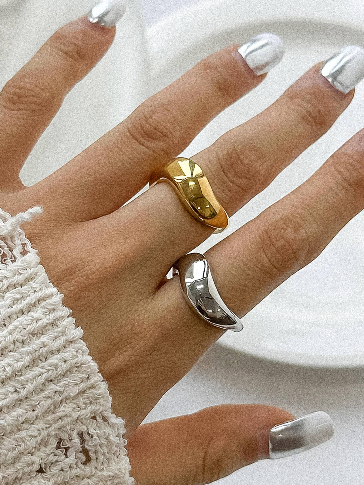 Hand wearing silver and gold sculptural rings with a smooth, minimalist design, paired with chic white nails for a trendy look