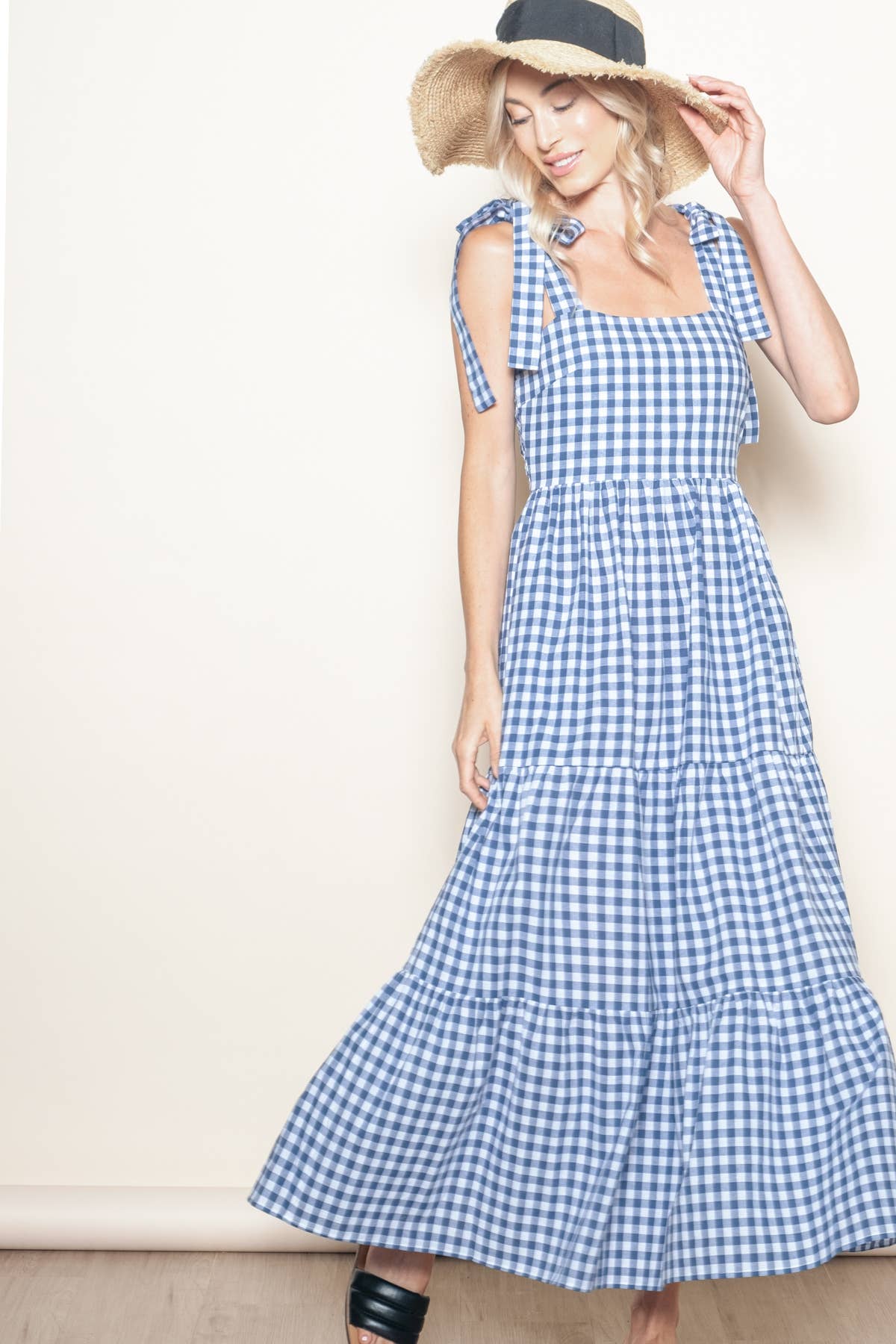  A blonde model wearing a blue and white gingham tiered maxi dress with tie straps, styled with a straw hat, posing in a graceful stance indoors against a light-colored background.