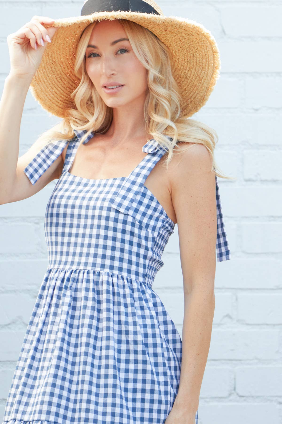 : A close-up of a blonde model in a blue and white gingham tiered maxi dress, accessorized with a straw hat, posing outdoors against a white brick wall.