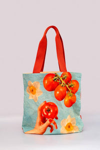 Canvas tote bag featuring a colorful design with ripe cherry tomatoes, daffodils, and a hand holding a tomato against a light blue background