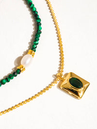 Close-up of a layered necklace featuring green beads, gold accents, a pearl detail, and a gold pendant with an emerald green stone.