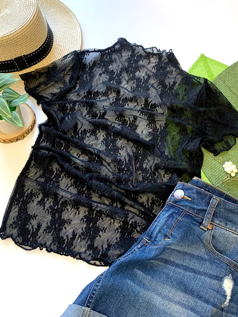 Flat lay of a black lace top with floral detailing, styled with denim jeans, a straw hat, and greenery for a stylish and versatile outfit