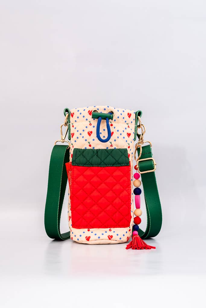 Colorful quilted water bottle sling with cherry print, green adjustable strap, red front pocket, and a playful beaded charm with a tassel detail.