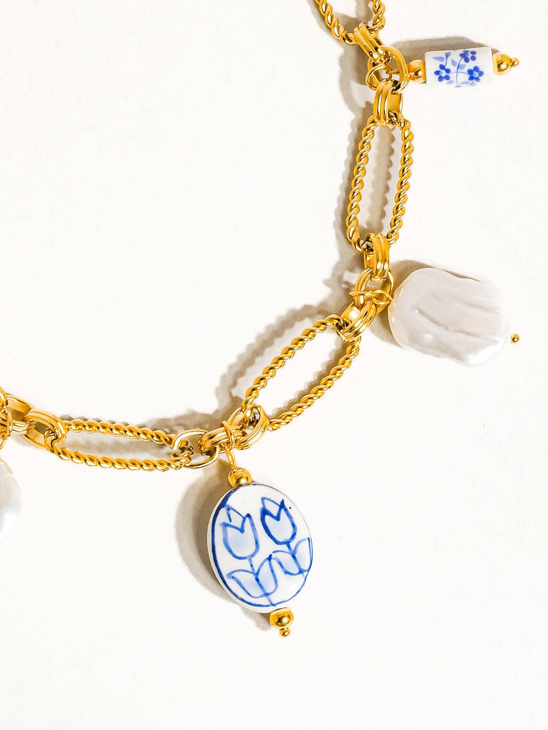 Close-up of a gold chain necklace featuring ceramic charms, including a blue tulip design, a white pearl-like charm, and gold link details