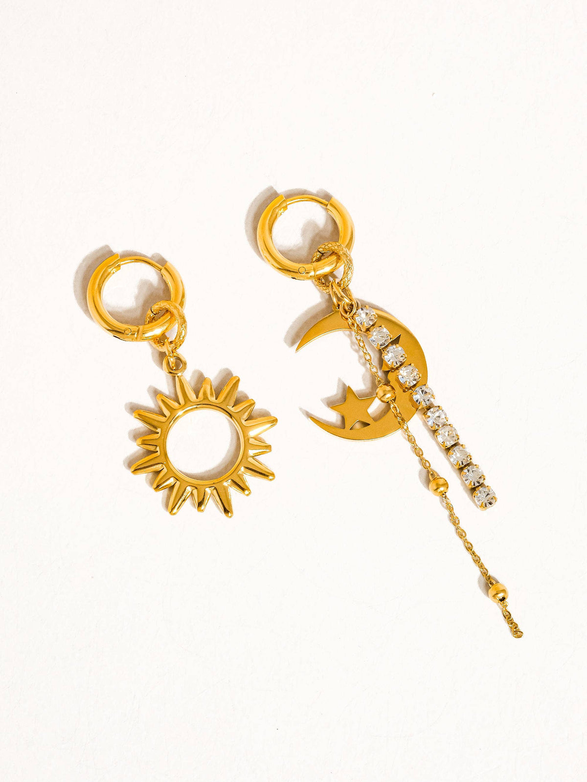 Flat lay of gold mismatched earrings featuring a sun charm and a crescent moon with dangling rhinestones, showcasing celestial-inspired jewelry