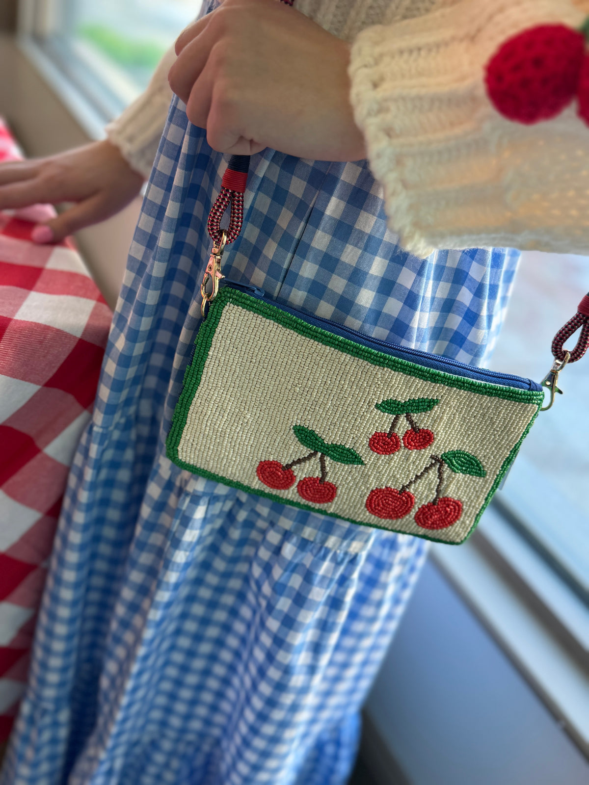 women holding cherry beaded cluth purse wearing a blue and white ginham maxi dress with her hand on a red and white tablecloth