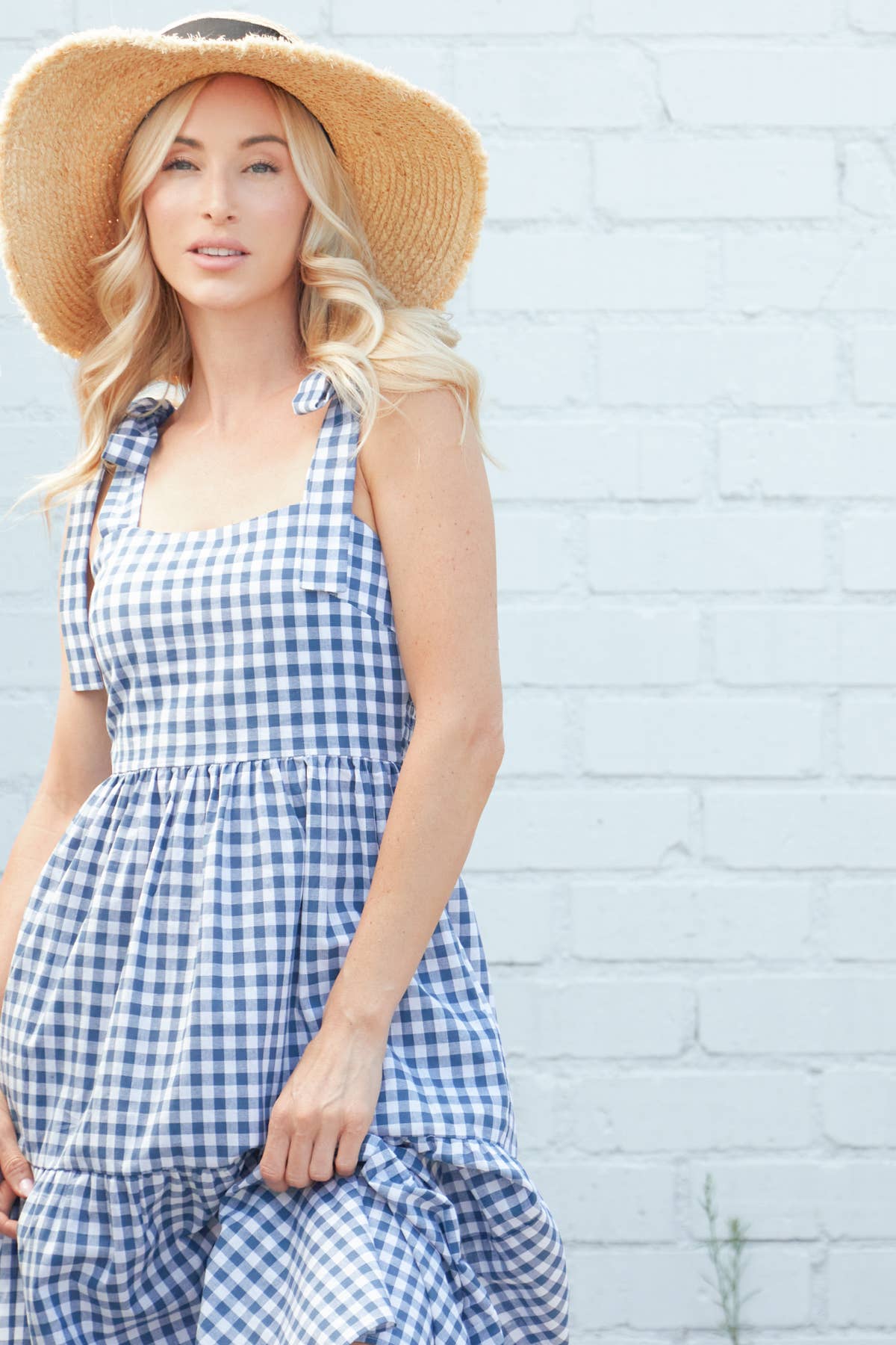 A blonde model in a blue and white gingham tiered maxi dress with tie straps, holding the skirt, styled with a straw hat, posing outdoors against a white brick wall.