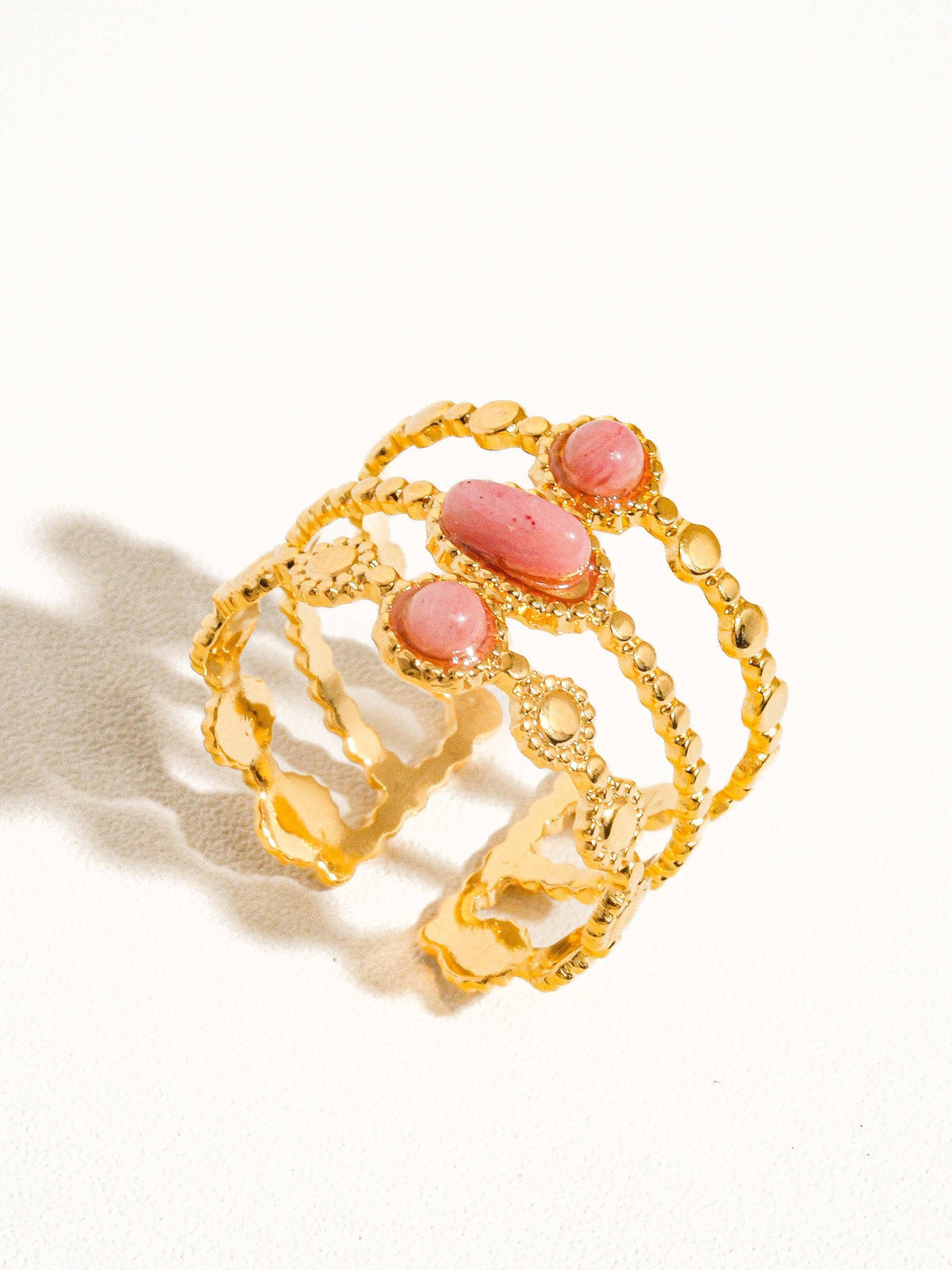 A gold-plated statement ring featuring three pink gemstone accents in a textured triple-band design, displayed on a white background.