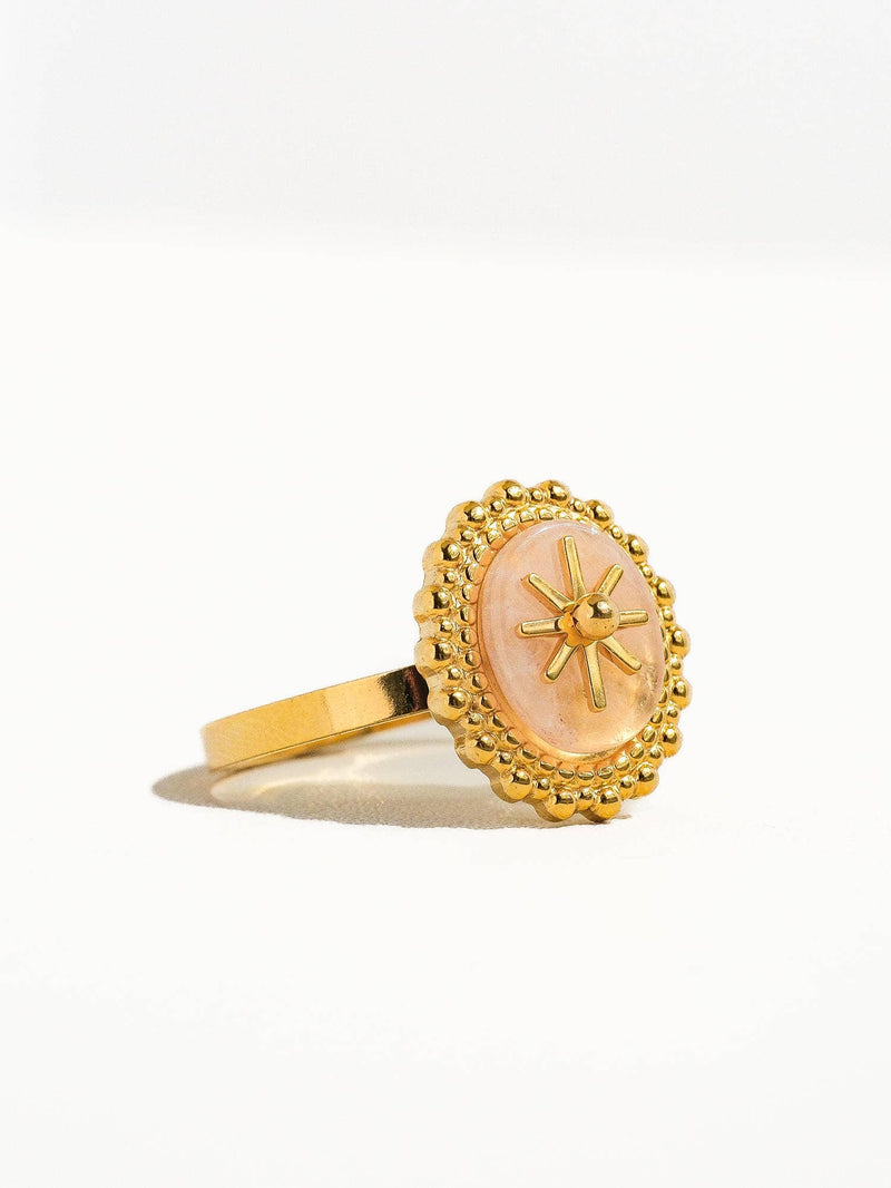 Angled view of a gold ring with a pink marbled oval stone, accented by a starburst detail and surrounded by beaded edging.