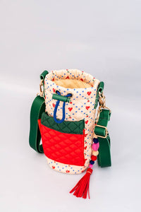 Colorful quilted water bottle sling with cherry print, green adjustable strap, red front pocket, and a playful beaded charm with a tassel detail.