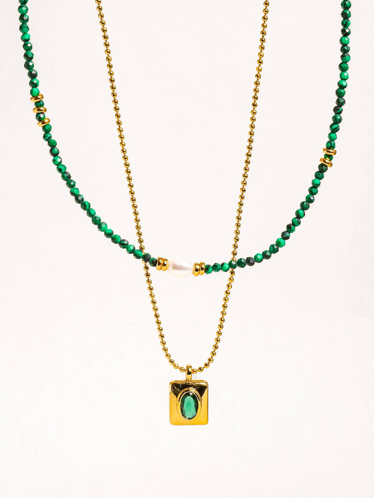 Close-up of a gold chain necklace with a rectangular pendant holding a green stone, paired with a green beaded layer and pearl accents