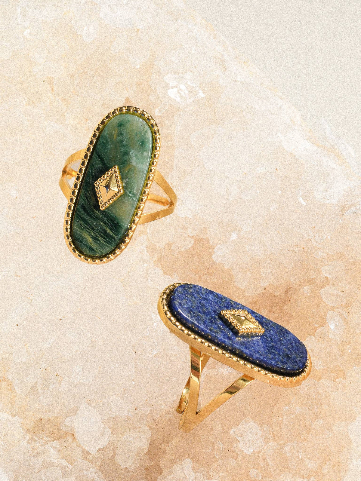 Two gold rings with oval-shaped stones, one green and one blue, both featuring intricate beaded borders and geometric accents, styled against a textured backdrop