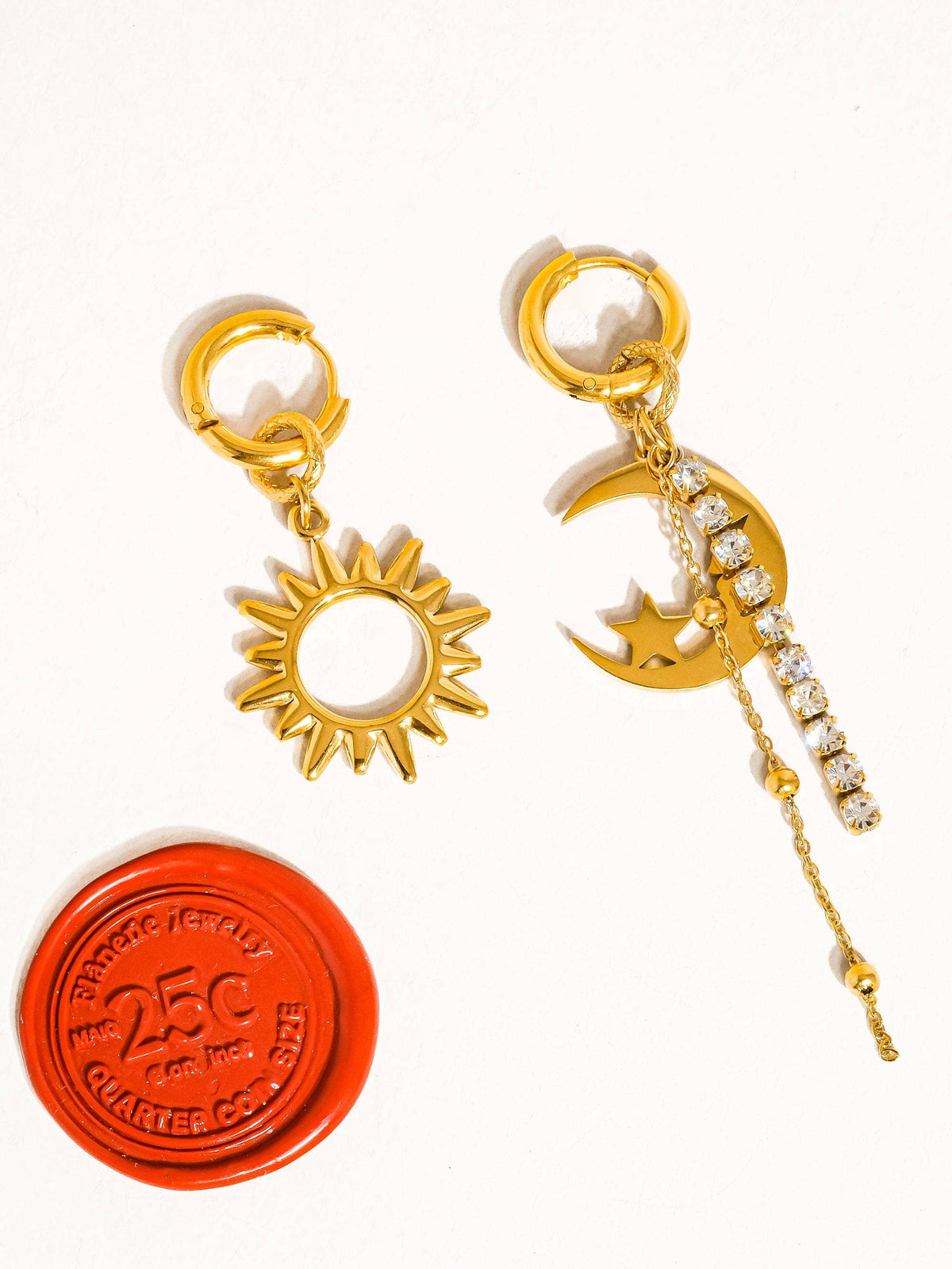 Pair of mismatched gold earrings featuring a sun charm and a crescent moon with dangling rhinestones and chains, displayed next to a red wax seal accent