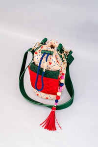 Colorful quilted water bottle sling with cherry print, green adjustable strap, red front pocket, and a playful beaded charm with a tassel detail.