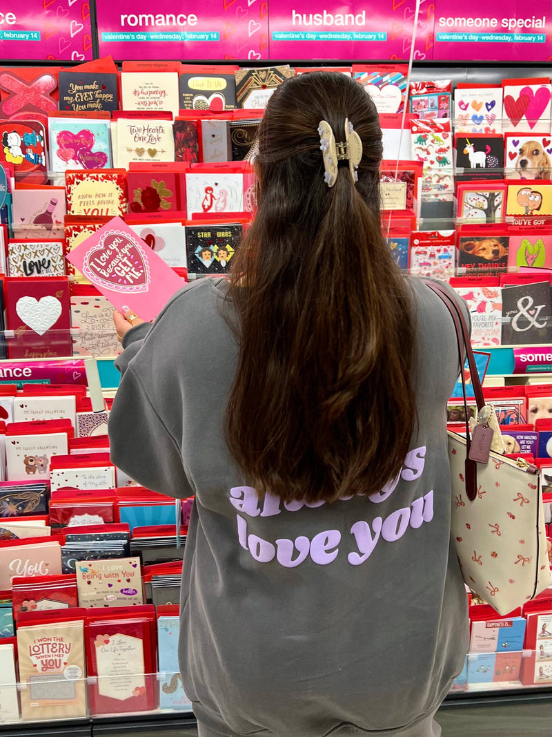 A person wearing the "I'll Always Love You" dark gray fleece sweatshirt, showcasing the oversized fit and lavender puff print text on the back. They are browsing a Valentine's Day card aisle, adding a sentimental touch to this cozy, sustainable loungewear piece.