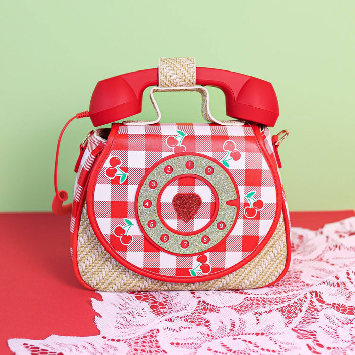 Close-up of the cherry-themed handbag with gingham print, glittery rotary dial, and structured shape on a lace and red backdrop.