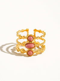 A side-angle view of a gold-plated ring with three pink gemstones, showing intricate detailing and textured bands, resting on a white surface.