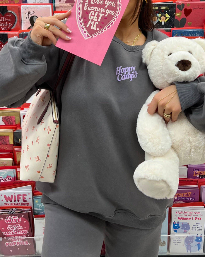 A person wearing the "I'll Always Love You" dark gray fleece sweatpants and matching sweatshirt featuring the "Happy Camp3r" lavender puff print logo. They are holding a Valentine's Day card and a plush teddy bear, standing in front of a display of greeting cards. Cozy, unisex, and made from sustainable materials, these sweatpants are perfect for everyday comfort and style.