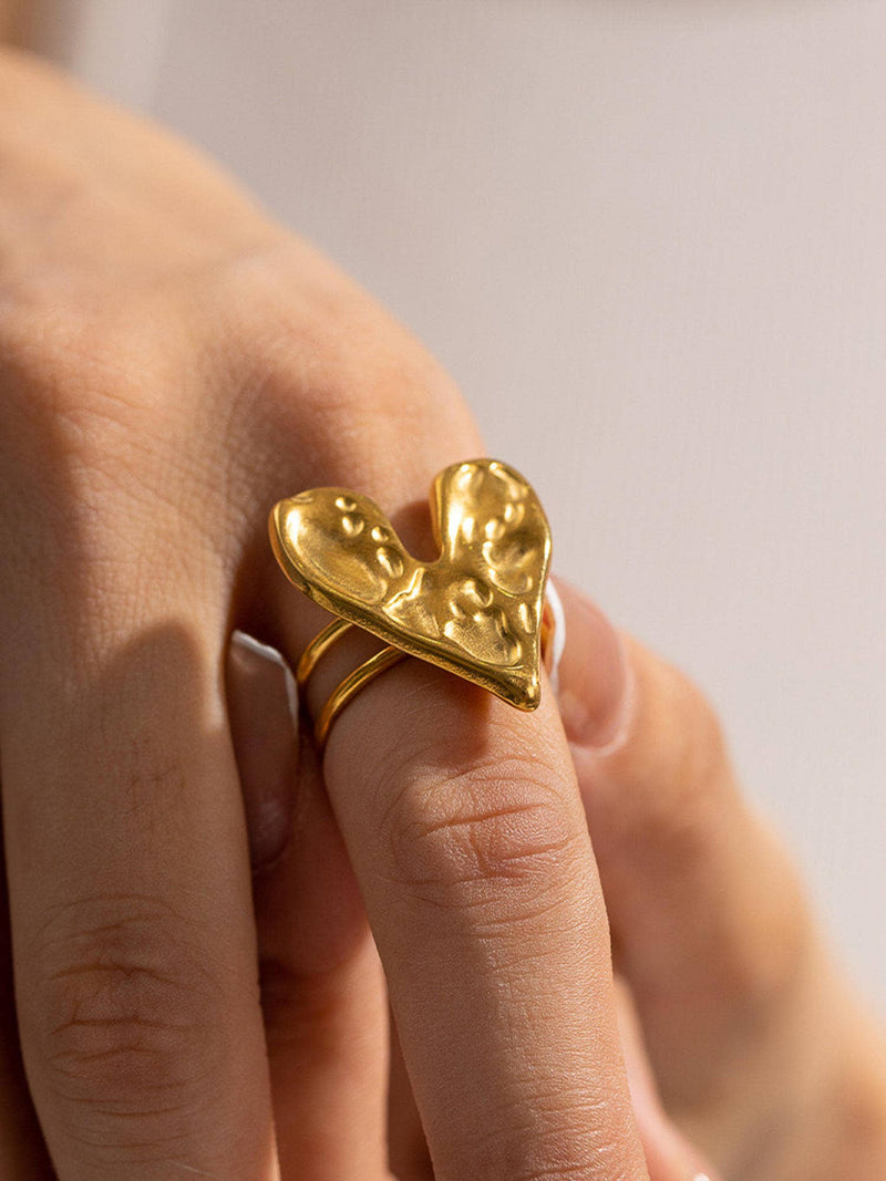 A close-up view of a gold ring featuring a sculptural, textured heart design worn on a finger, showcasing its bold and artistic style.