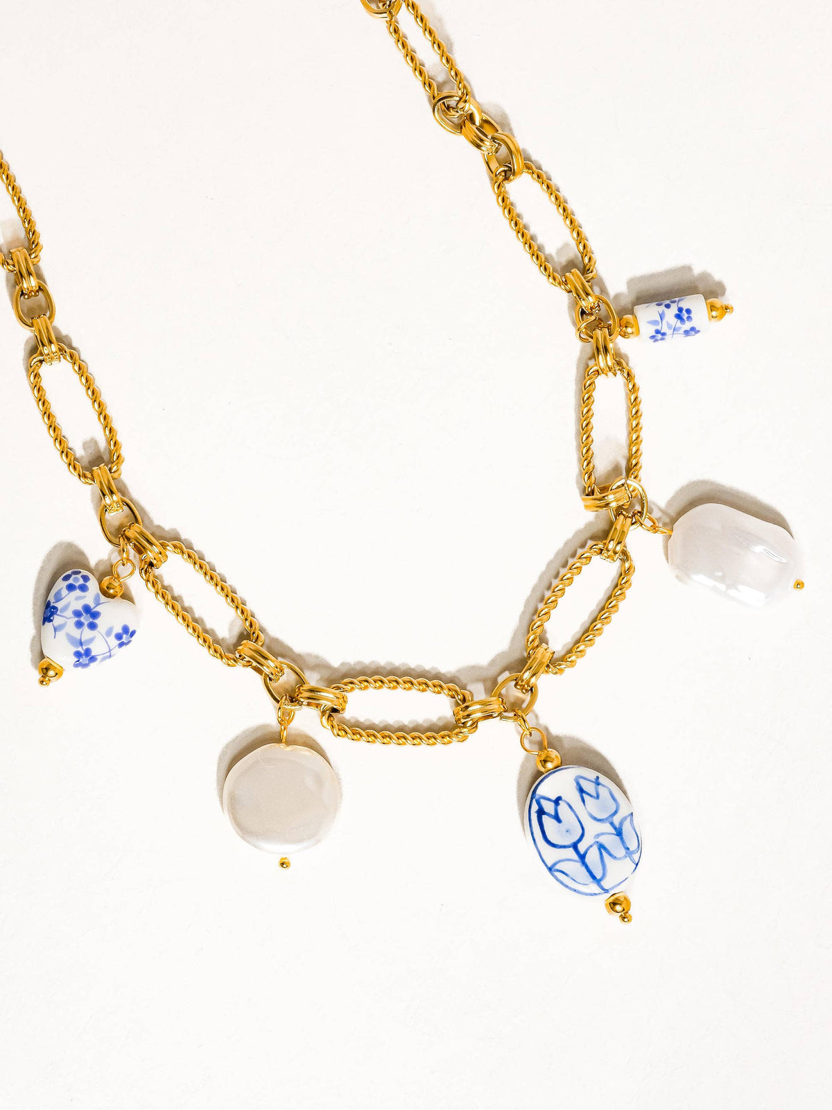 Gold chain necklace with ceramic charms, including blue floral patterns and white accents, displayed flat to showcase its design.