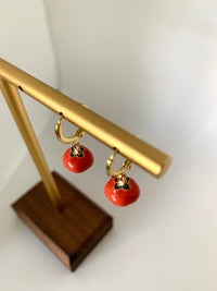 Gold-tone hoop earrings with small red tomato charms hanging from a jewelry display stand.