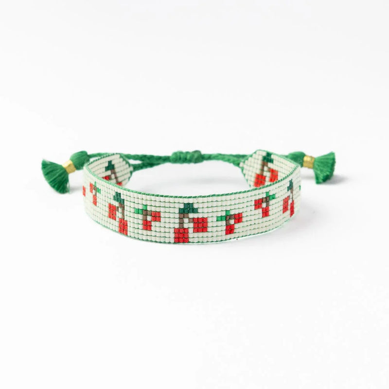 A handwoven beaded bracelet with a cherry pattern on a cream background, featuring green adjustable braided ties and tassel ends for a playful, fruity touch.